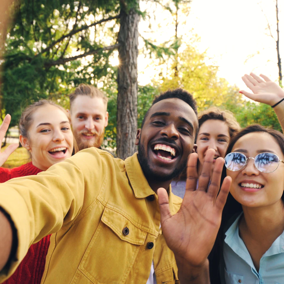 A group of young friends laughing and waving together outdoors in a sunny green park