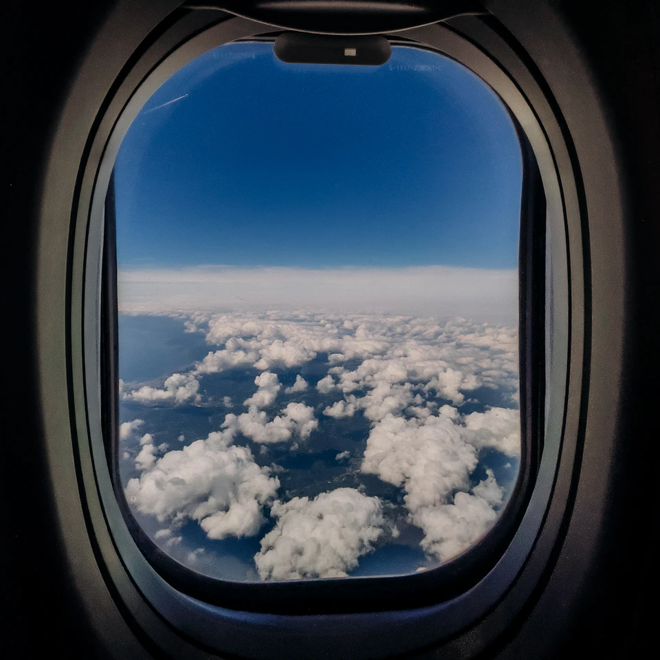 An airplane window view of bright blue sky and fluffy white clouds