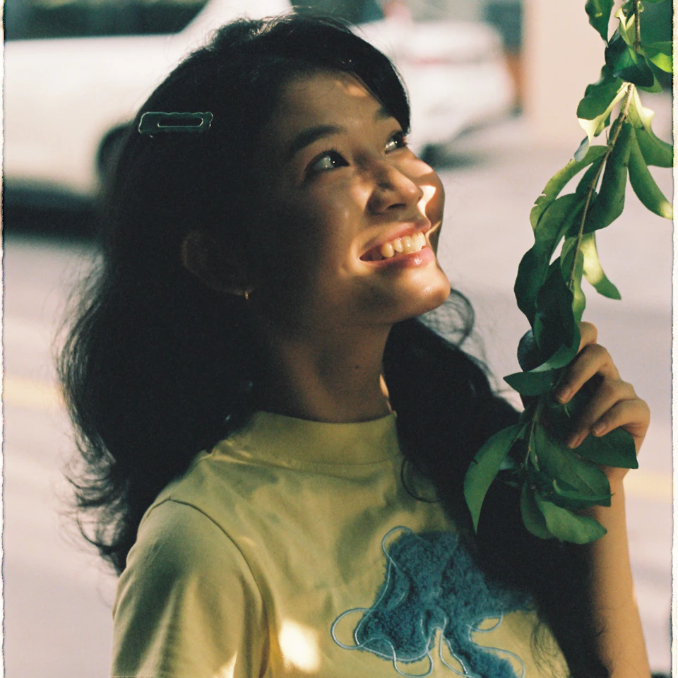 A young woman smiling softly as she looks up toward fresh green leaves in bright daylight