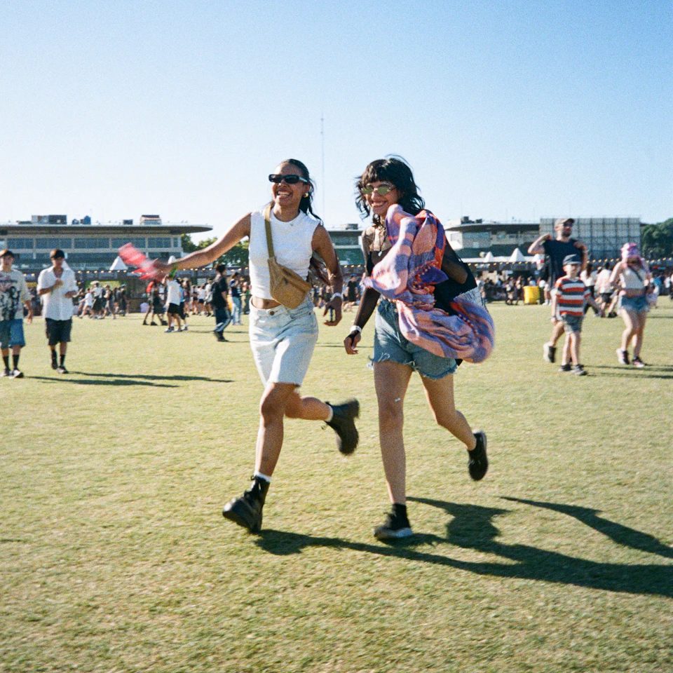 Two young women running and laughing across a sunny open field under a bright blue sky