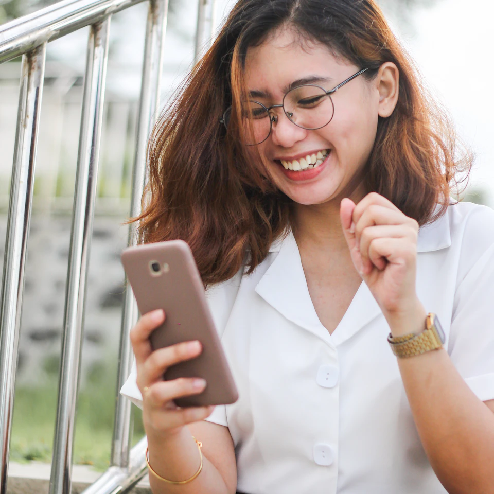 A young woman laughing while looking at travel photos on her phone in bright daylight