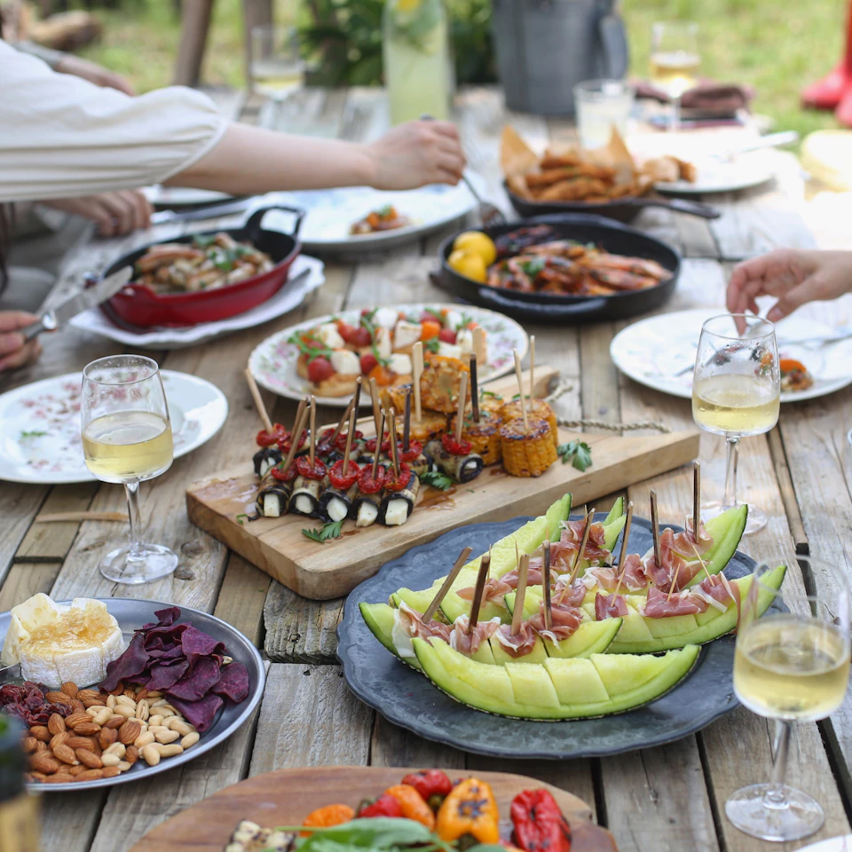 Friends sharing a colorful outdoor meal around a sunny wooden table in bright daylight