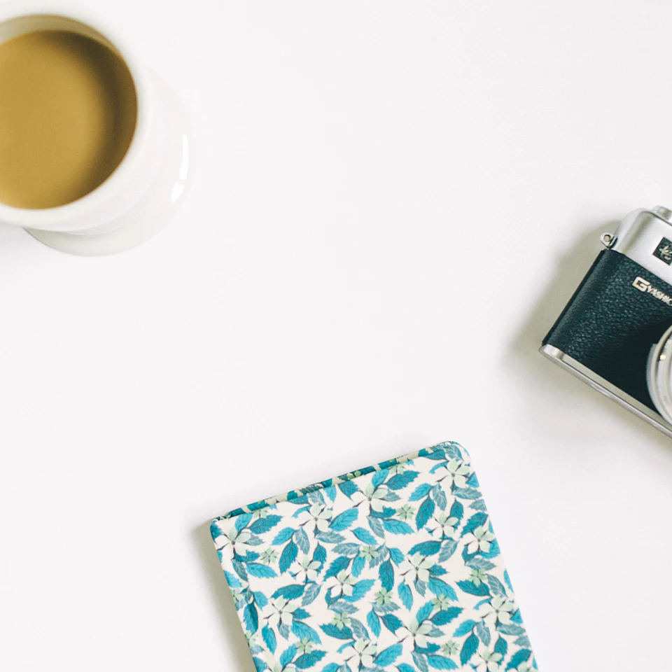 A bright airy flat lay of a vintage film camera, a floral notebook and coffee on a white desk